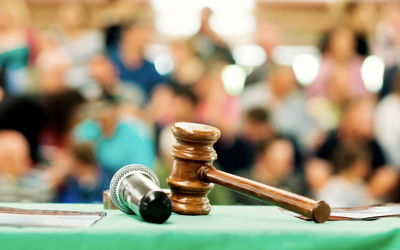 Eine Szene aus einer Versteigerung, bzw. Auktion. Ein Auktionshammer und ein Mikro liegen vor einer Menschengruppe.