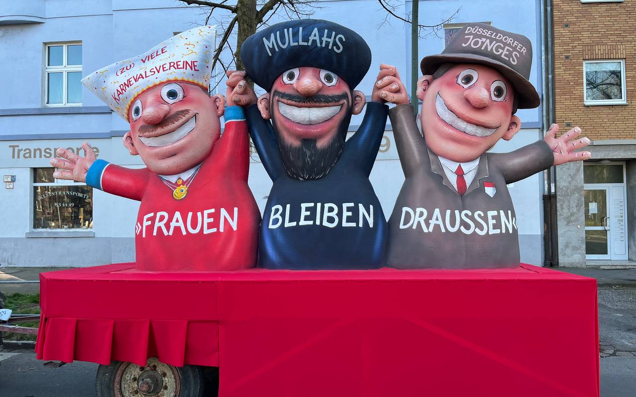 Mottowagen beim Karneval:  Karnevalsvereine, Mullahs und die Düsseldorfer Jonges halten sich an den Händen und reißen die Arme hoch. Vorne steht "Frauen müssen draußen bleiben".