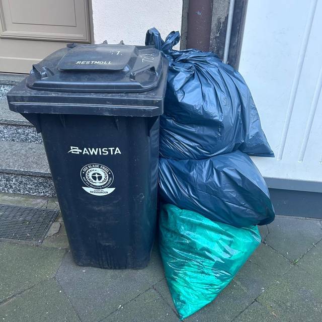 Eine Restmülltonne steht vor einer Hauswand, daneben mehrere verschlossene Säcke mit Restmüll.