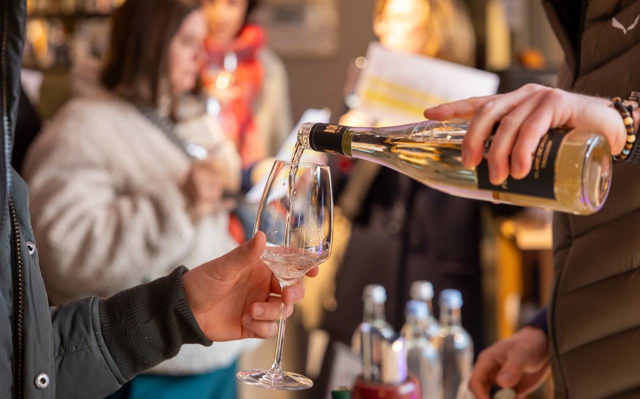 Ein Kellner schüttet Weißwein aus einer Flasche in ein Weinglas. Im Hintergrund sind verschwommen Wasserflaschen und Menschen zu sehen.