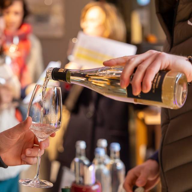 Ein Kellner schüttet Weißwein aus einer Flasche in ein Weinglas. Im Hintergrund sind verschwommen Wasserflaschen und Menschen zu sehen.