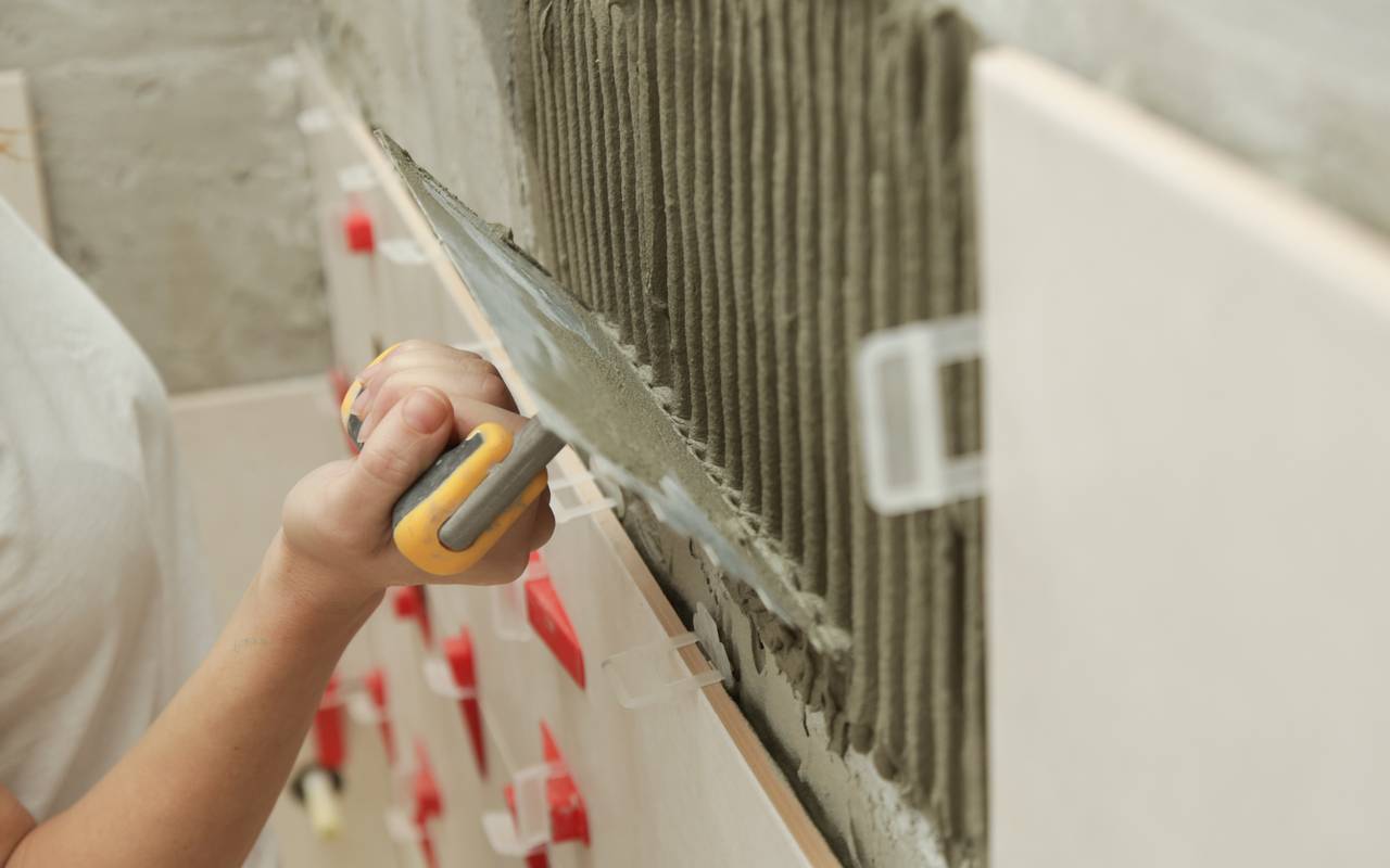 Frau beim Fliesen der Badezimmerwände, die Klebstoff auf eine Fliese aufträgt und die Keramikfliese in die richtige Position bringt.