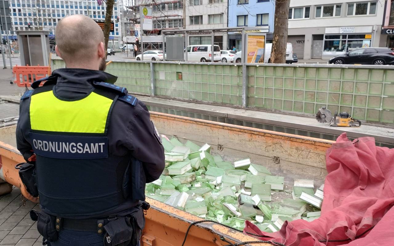 Ein Mitarbeiter des Ordnungsamtes steht vor einem Container mit herausgeschlagenen Glasbausteinen. Im Hintergrund die grünen Bänke am Worringer Platz, die jetzt nach und nach entfernt werden.