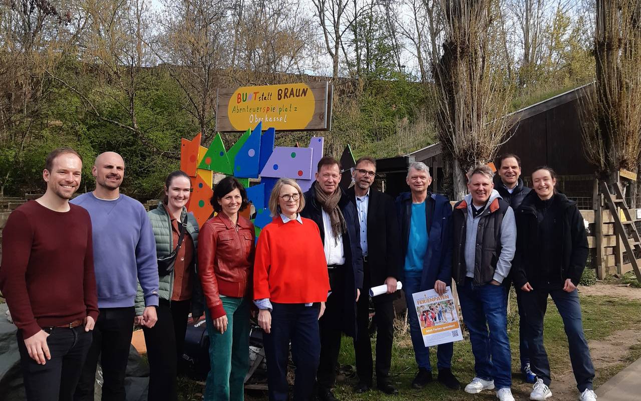 Ein Menschengruppe hat sich vor einem bunten Schild aufgestellt, alle Lächeln in die Kamera. Im Hintergrund sind Bäume in Sträucher.