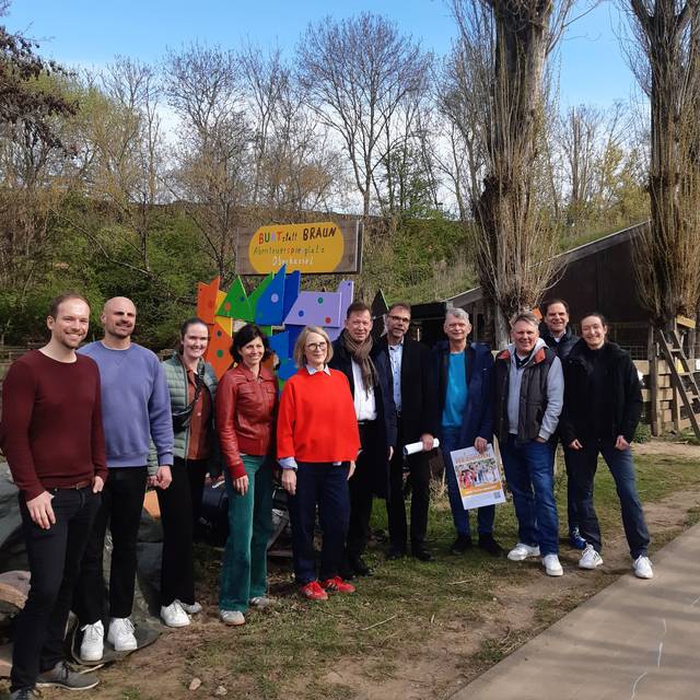 Ein Menschengruppe hat sich vor einem bunten Schild aufgestellt, alle Lächeln in die Kamera. Im Hintergrund sind Bäume in Sträucher.