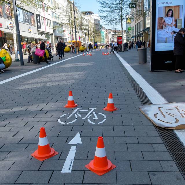Der Radweg auf der Schadowstraße ist mit weißer Farbe teilweise neu markiert. Zu sehen ist unter anderem ein Fahrrad-Piktogramm. Links und rechts vor den Geschäftshäusern sind Menschen zu Fuß unterwegs.