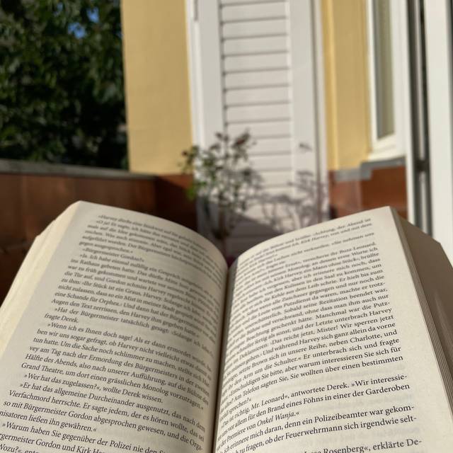 Ein aufgeschlagenes Buch. Im Hintergrund ist ein Balkon zusehen, mit einer weißen Tür und einer gelben Wand.