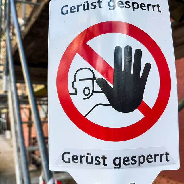 An einem Gerüst hängt ein Schild, auf dem ein durchgestrichener Roter Kreis, ein Mensch und der Schriftzug "Gerüst gesperrt" zu sehen sind.