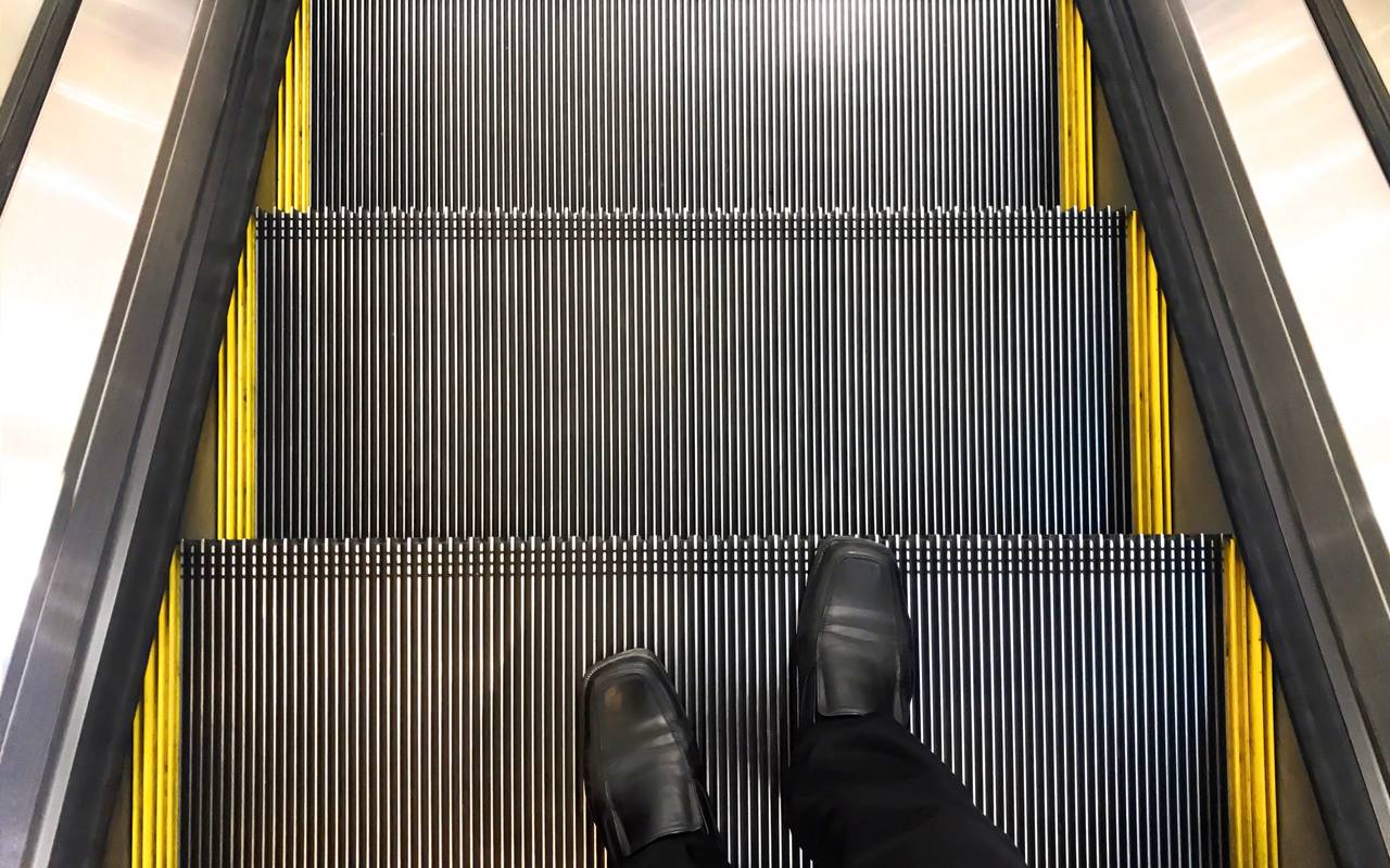 Ein Geschäftsmann mit schwarzen Lederschuhen. Er fährt eine Rolltreppe hinunter.