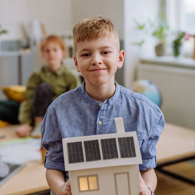 Ein Junge hält ein gebasteltes Holzhaus mit Solaranlagen auf dem Dach in die Kamera und lächelt. Im Hintergrund weitere Schulkinder, die an einem Tisch an etwas arbeiten.