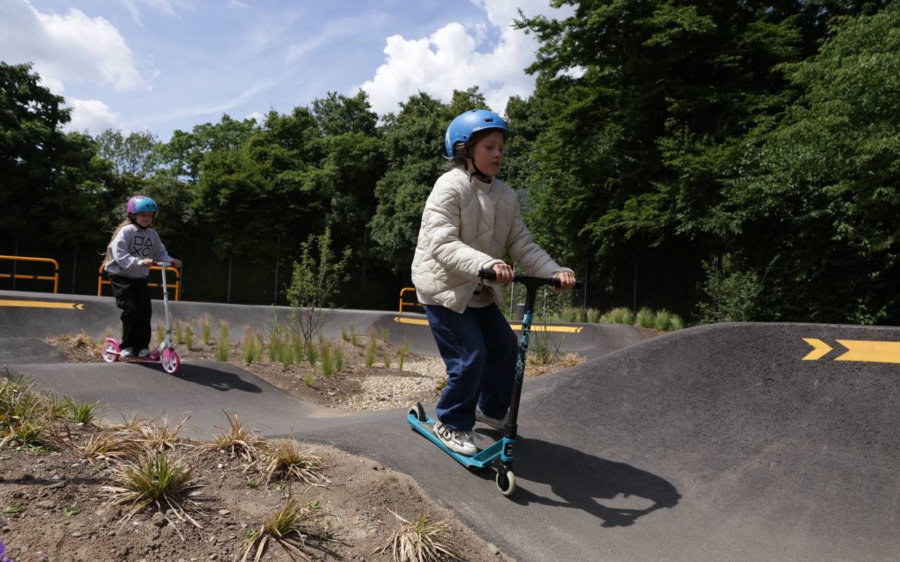 Zwei Kinder mit Helm testen auf ihren Rollern die neue Pumptrackanlage in Heerdt. Im Hintergrund befinden sich mehrere Bäume auf der Anlage.