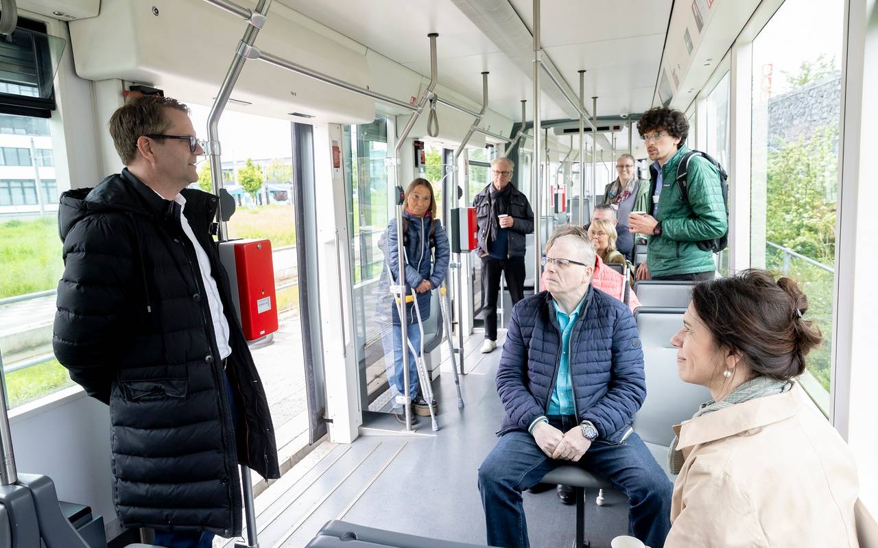 Mehrere Menschen sitzen und stehen im Inneren einer Straßenbahn. In der Mitte steht ein Mann und spricht, die anderen schauen ihn an.