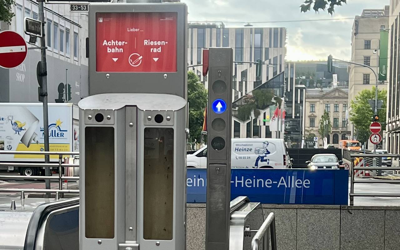 Vor dem Abgang zum U-Bahnhof "Heinrich-Heine-Allee" steht der neue Abstimm-Aschenbecher mit der Frage "Achterbahn oder Riesenrad?". Im Hintergrund ist unter anderem der Kö-Bogen zu sehen.