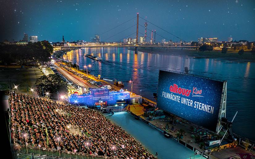 Eine Tribüne voller Menschen steht vor einer Kinoleinwand am Rhein, im Hintergrund der Rheinturm.
