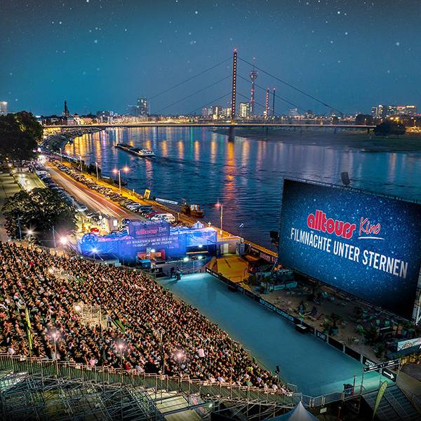 Eine Tribüne voller Menschen steht vor einer Kinoleinwand am Rhein, im Hintergrund der Rheinturm.