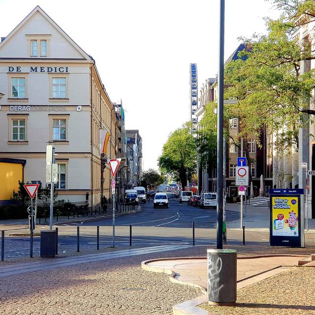 Ein städtischer Straßenzug mit einem gelb-weißen Gebäude, geparkten Autos und einem Riesenrad im Hintergrund; rechts ein Backsteingebäude mit Werbetafel.