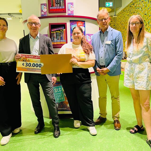 Fünf Menschen stehen in der Stadtbücherei und freuen sich über einen Gutschein über 30.000 Euro der Deutschen Postcode Lotterie
