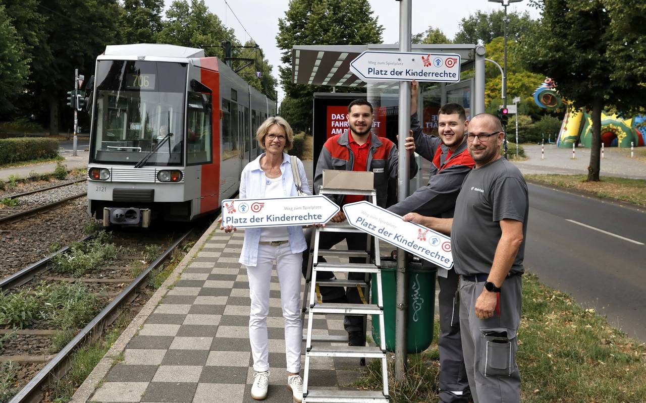 Mitarbeitende von Rheinbahn und Kinderschutzbund mit Schildern (den Platz der) Kinderrechte. Sie stehen neben einer Bahn an der Haltestelle "Volksgarten S".