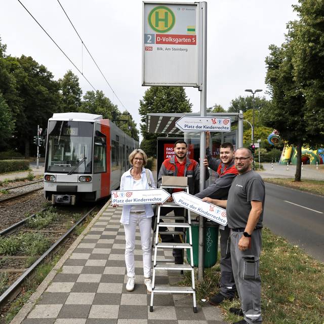 Mitarbeitende von Rheinbahn und Kinderschutzbund mit Schildern (den Platz der) Kinderrechte. Sie stehen neben einer Bahn an der Haltestelle "Volksgarten S".