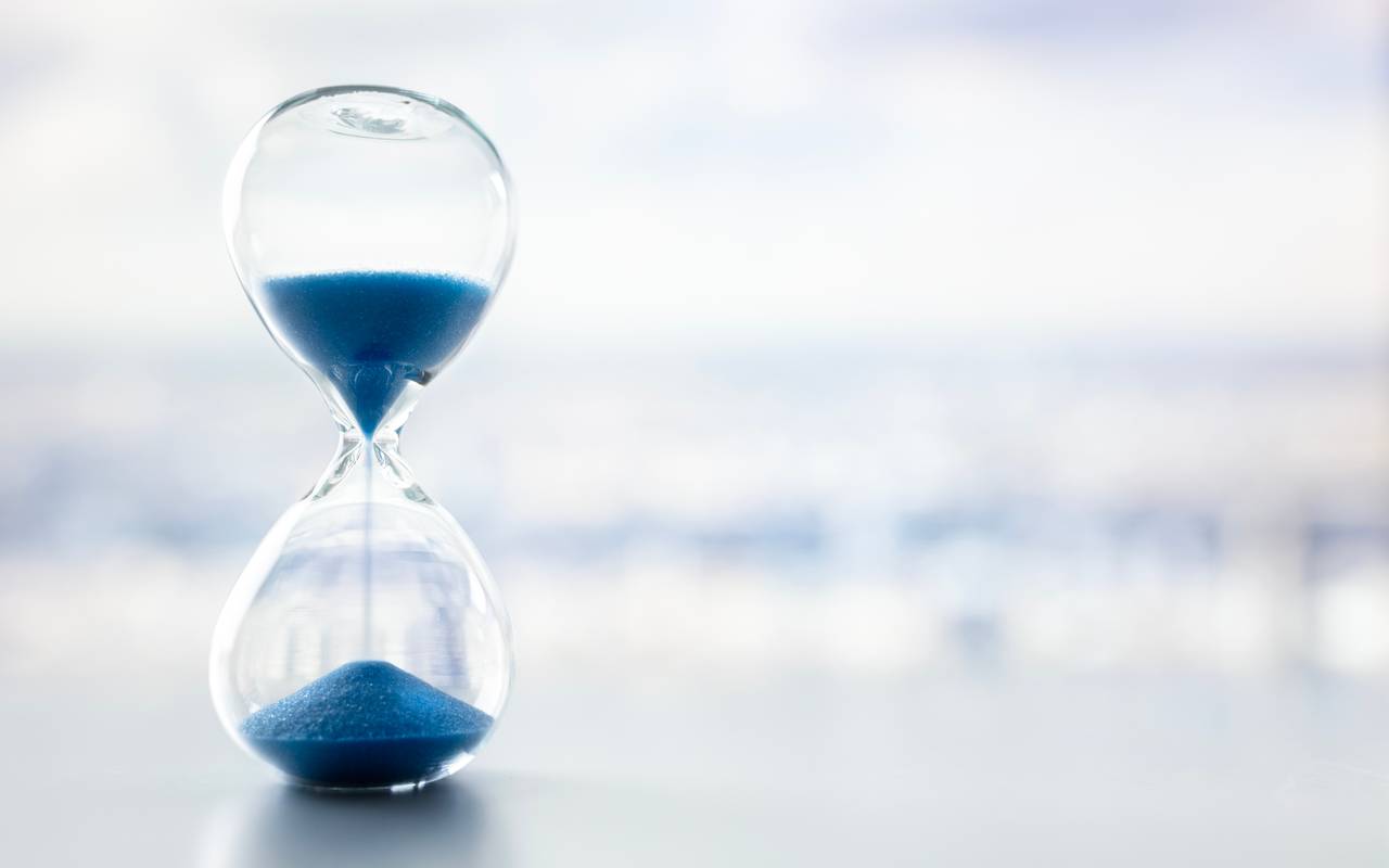 Eine Sanduhr mit blauem Sand vor einem grau-weißen milchigen Hintergrund.