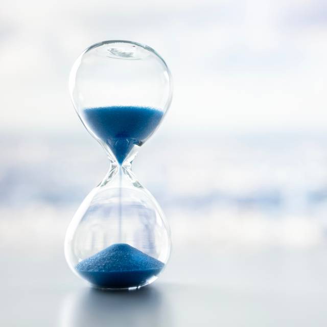 Eine Sanduhr mit blauem Sand vor einem grau-weißen milchigen Hintergrund.