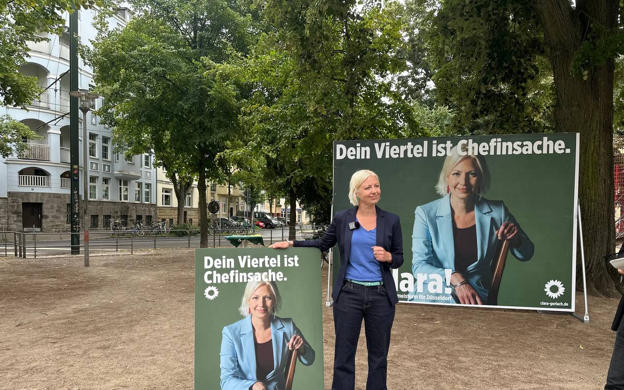 Oberbürgermeister-Kandidatin Clara Gerlach steht zwischen ihren Wahlplakaten, auf denen jeweils Clara Gerlach zu sehen ist. Links ein Laternenplakat, rechts ein Großplakat.