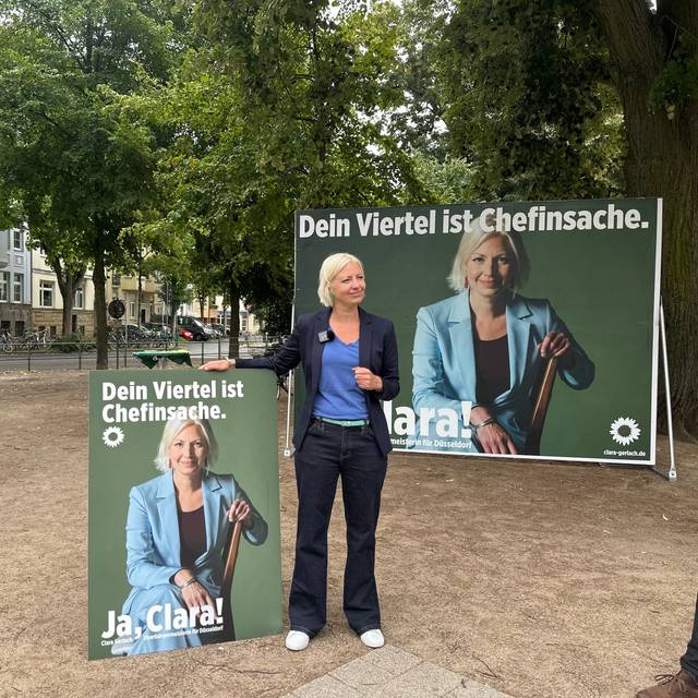 Oberbürgermeister-Kandidatin Clara Gerlach steht zwischen ihren Wahlplakaten, auf denen jeweils Clara Gerlach zu sehen ist. Links ein Laternenplakat, rechts ein Großplakat.