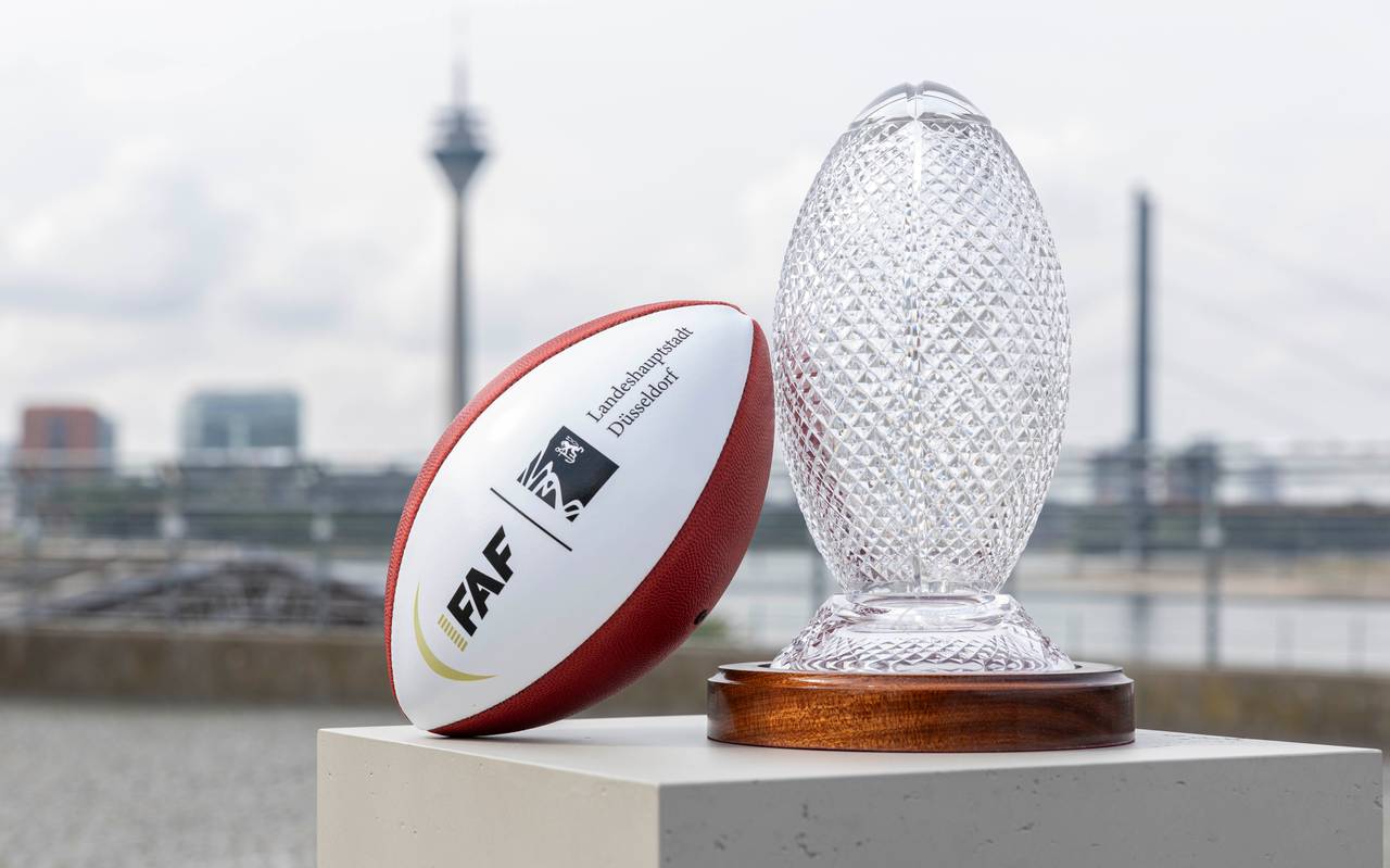 Ein Football und der Pokal der Flag Football WM stehen auf dem Burgplatz. Im Hintergrund sind die Rheinkniebrücke und der Rheinturm.