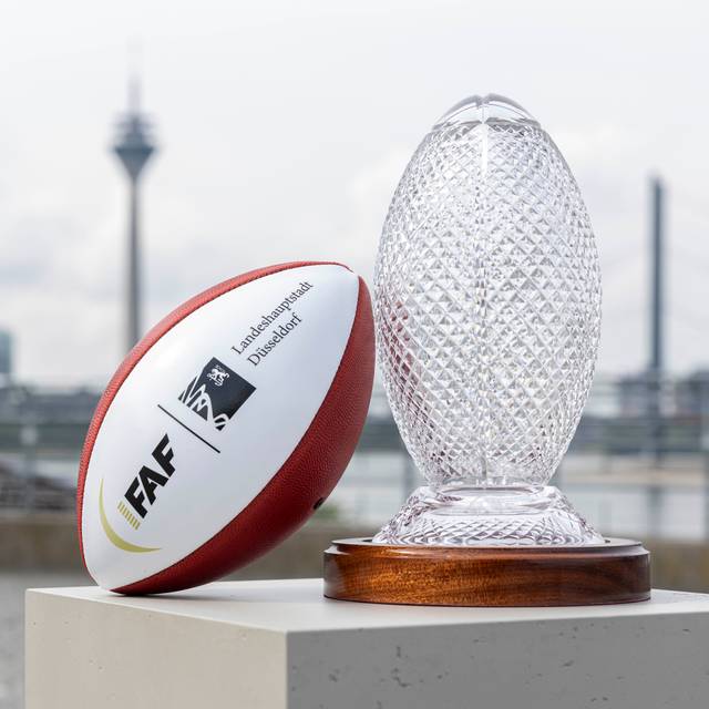 Ein Football und der Pokal der Flag Football WM stehen auf dem Burgplatz. Im Hintergrund sind die Rheinkniebrücke und der Rheinturm.