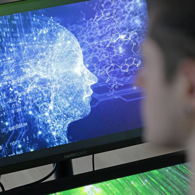 Eine Person sitzt vor einem Bildschirm, auf dem zu erkennen ist, dass es neben dem menschlichen Kopf auch verschiedene Ströme gibt, die fließen. Eine Art Symbolbild für Künstliche Intelligenz