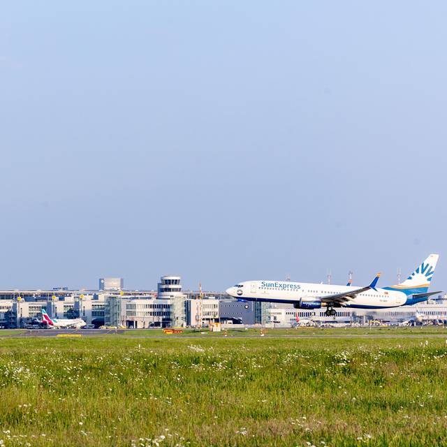 Rechts im Bild ein Flugzeug auf der Startbahn, das gerade abhebt, davor Rasen. Im Hintergrund der Düsseldorfer Flughafen, davor stehen einige Flugzeuge.