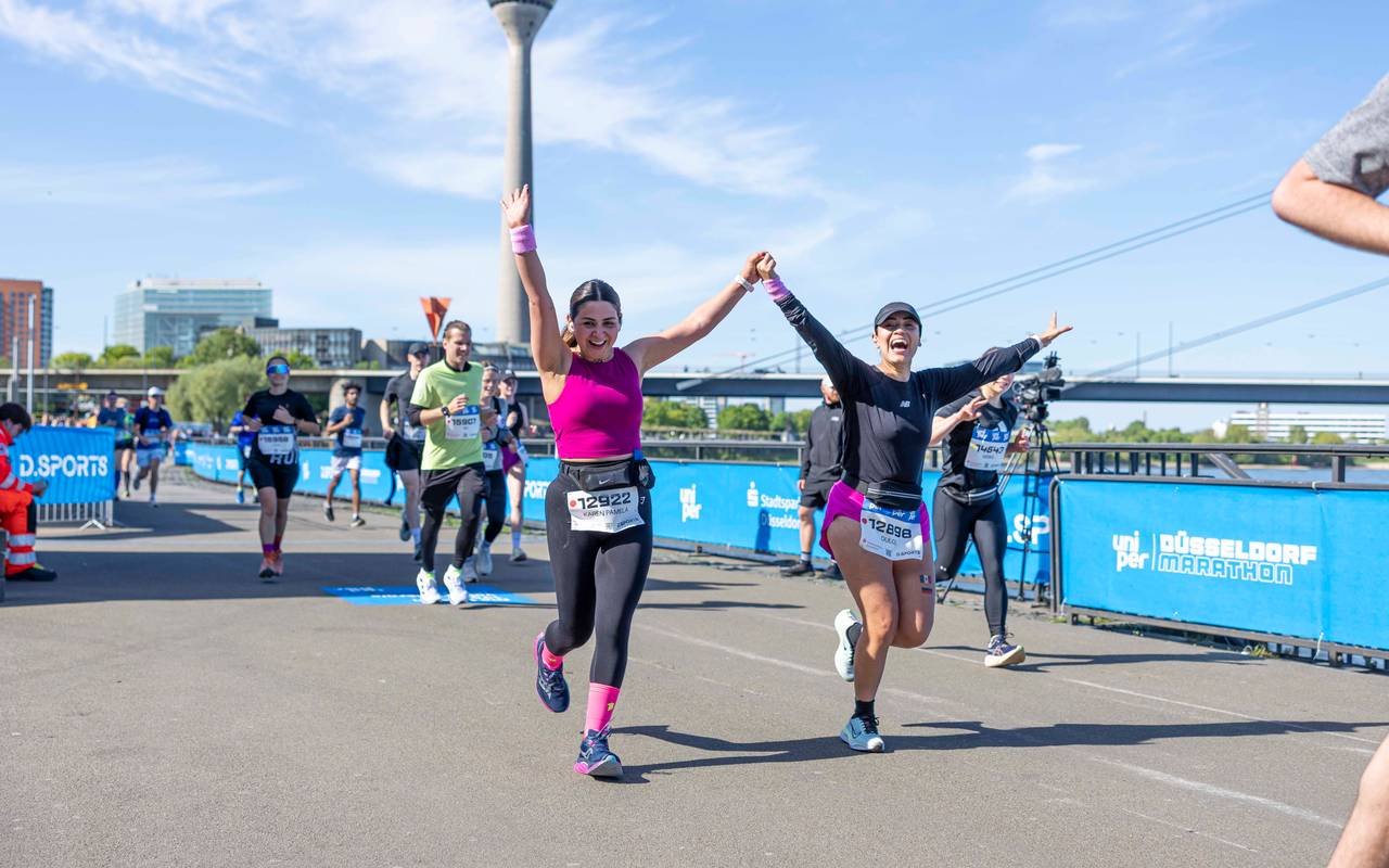Zwei Frauen laufen beim Düsseldorf Marathon am Rheinufer entlang und reißen freudig die Arme hoch. Im Hintergrund der Rheinturm.