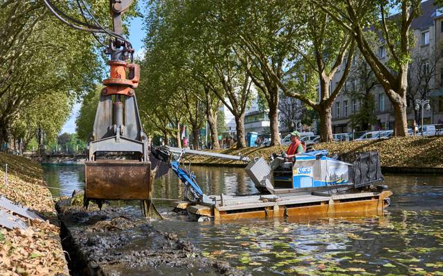 Ein Amphibienfahrzeug steht im Wasser eines Grabens und schiebt Schlamm an den Rand. Ein Bagger hebt den Schlamm heraus.