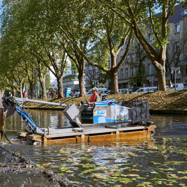 Ein Amphibienfahrzeug steht im Wasser eines Grabens und schiebt Schlamm an den Rand. Ein Bagger hebt den Schlamm heraus.