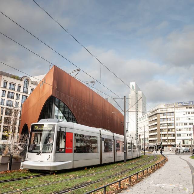 Eine Straßenbahn der Düsseldorfer Straßenbahn fährt durch das Stadtzentrum von Düsseldorf. Die Bahn wird von der Rheinbahn betrieben und ist Teil des öffentlichen Nahverkehrs. Im Hintergrund sind städtische Gebäude und Passanten zu sehen.