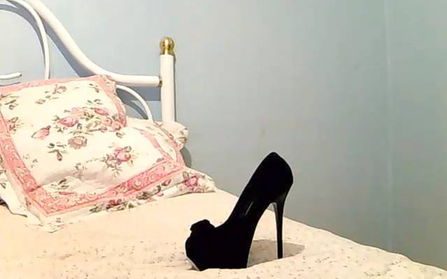 Schwarze Highheels auf einem Bett. Zu sehen ist auch ein rosa Kissen.