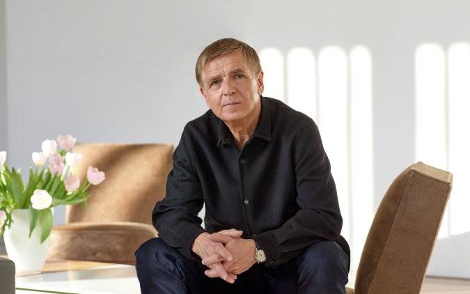 Ein Mann Sitzt auf einem Sessel und blickt in die Kamera. Er stützt seine Ellenbogen auf seinen Knien ab und faltet seine Hände ineinander.