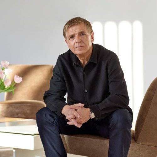 Ein Mann Sitzt auf einem Sessel und blickt in die Kamera. Er stützt seine Ellenbogen auf seinen Knien ab und faltet seine Hände ineinander.