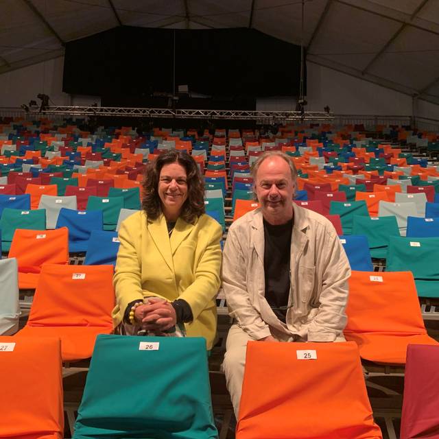 Eine Tribüne mit bunten Stühlen, von unten fotografiert. In der zweiten Reihe sitzen eine Frau und ein Mann und lächeln in die Kamera.