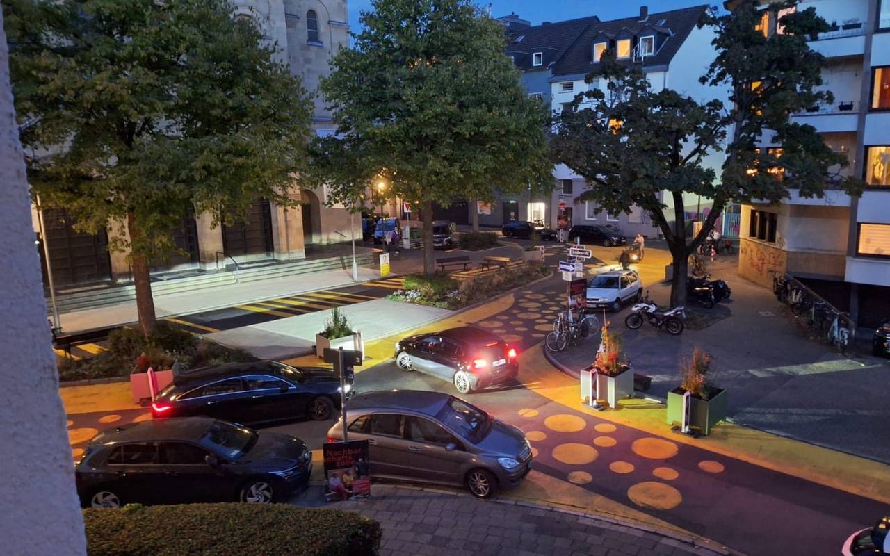 Auf dem Bild sieht man den Paulusplatz mit gelben Streifen und Punkten auf der Straße. Zusätzlich hat die Stadt Blumenkübel und Warnbaken aufgestellt. Mehrere Autos versuchen an den Kübeln vorbeizufahren.