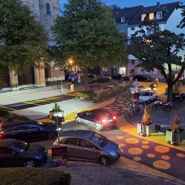 Auf dem Bild sieht man den Paulusplatz mit gelben Streifen und Punkten auf der Straße. Zusätzlich hat die Stadt Blumenkübel und Warnbaken aufgestellt. Mehrere Autos versuchen an den Kübeln vorbeizufahren.