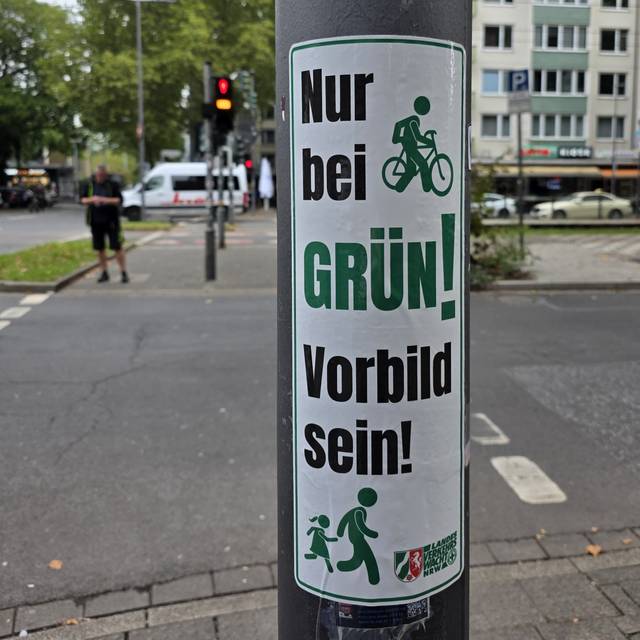 Ein grün-weißer Aufkleber mit dem Slogan "Nur bei Grün! – Vorbild sein!" klebt auf einem Ampelmast. Im Hintergrund ist eine Kreuzung zu sehen.