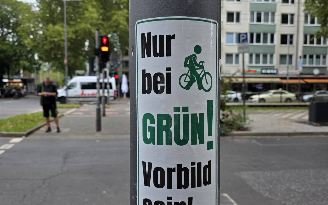 Ein grün-weißer Aufkleber mit dem Slogan "Nur bei Grün! – Vorbild sein!" klebt auf einem Ampelmast. Im Hintergrund ist eine Kreuzung zu sehen.