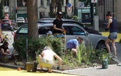 Mehrere Anwohnerinnen und Anwohner der Paulusstraße bepflanzen ein Beet um einen Baum herum. Im Hintergrund sind parkende Autos und die Straße sowie eine rote Ampel.