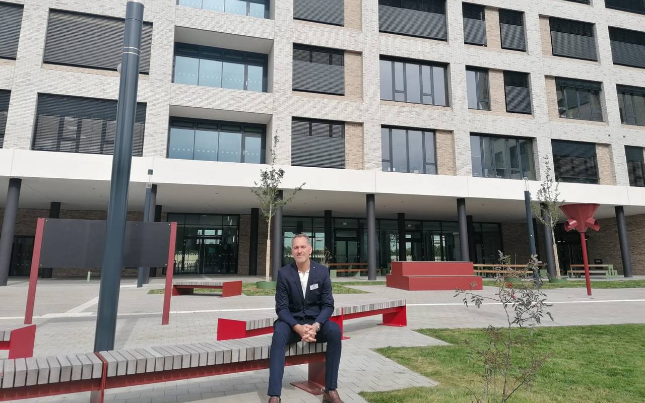 Schulleiter Jens Kock sitzt auf dem Schulhof auf einer Bank und blickt in die Kamera. Hinter ihm der Neubau der Realschule Benrath.