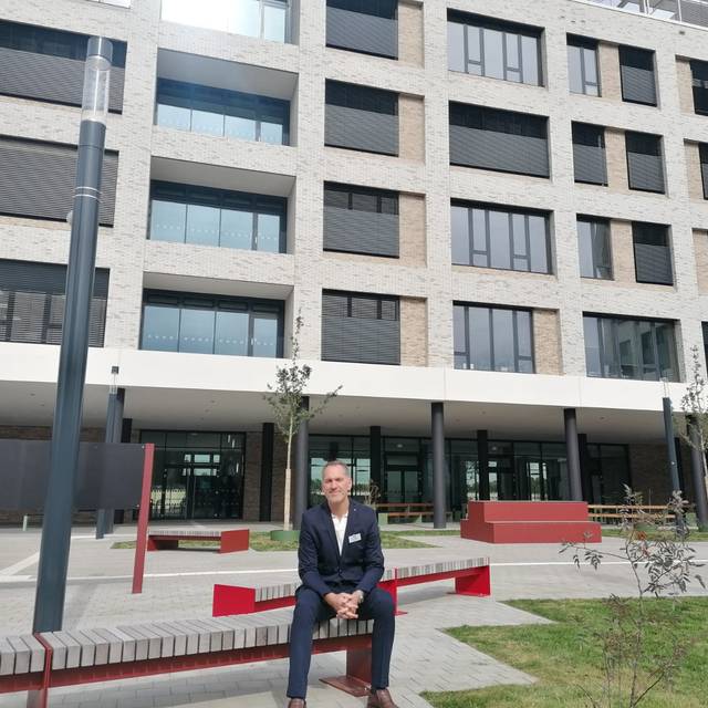 Schulleiter Jens Kock sitzt auf dem Schulhof auf einer Bank und blickt in die Kamera. Hinter ihm der Neubau der Realschule Benrath.