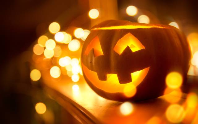 Ein fröhlich lächelnder Jack O’Lantern steht zu Halloween auf einem Kamin, umgeben von Lichterketten.
