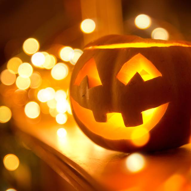 Ein fröhlich lächelnder Jack O’Lantern steht zu Halloween auf einem Kamin, umgeben von Lichterketten.