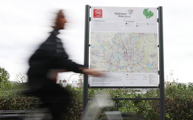 Vor einem Schild mit einer großen Fahrradkarte von Düsseldorf und der Region fährt ein Radfahrer entlang. Im Hintergrund sind der Schlossturm und das Riesenrad zu erkennen.