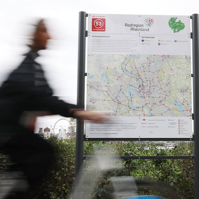 Vor einem Schild mit einer großen Fahrradkarte von Düsseldorf und der Region fährt ein Radfahrer entlang. Im Hintergrund sind der Schlossturm und das Riesenrad zu erkennen.
